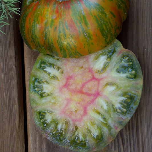 tomato sliced in half on a wood plank background. The skin is red with green, orange, and bronze striping, while the inside flesh is green with yellow and red marbling in the middle and some red streaks throughout the edges. 