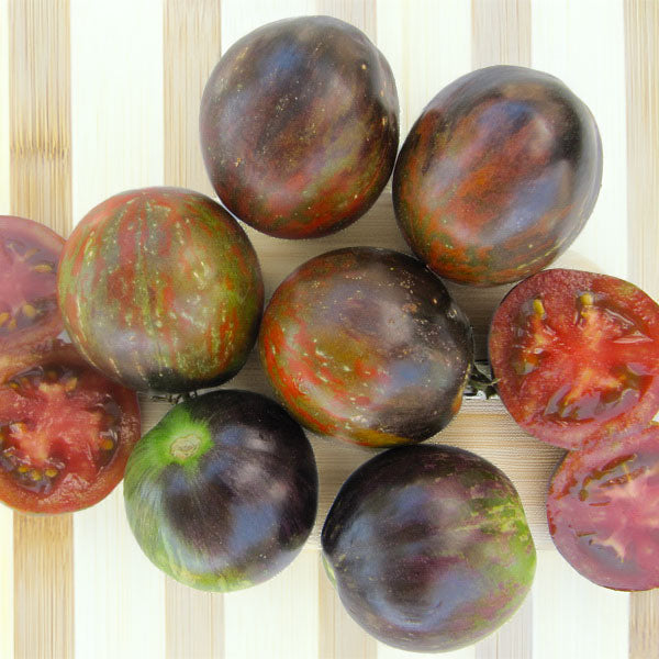 6 whole tomatoes on a bamboo cutting board. 2 unripe are green and dark purple (almost black) . 4 of them are half red, half dark purple with streaks and speckles of greens, 2 tomatoes are cut in half to reveal a deep red flesh